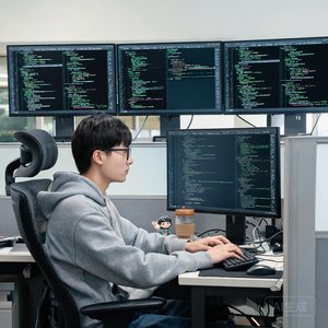 一位男性程序员在工位上，身后是多屏幕代码界面